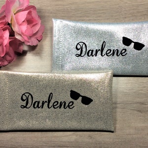 Pu&ograve; includere: Due custodie per occhiali argentate con il nome "Darlene" in corsivo e una grafica di occhiali da sole neri. Le custodie hanno una finitura strutturata e scintillante. Rose rosa sullo sfondo. Le custodie sono rettangolari.