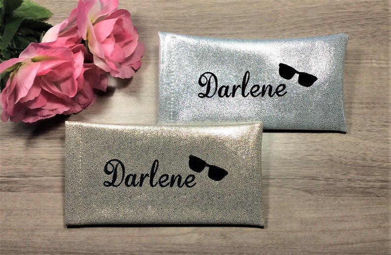 Pu&ograve; includere: Due custodie per occhiali argentate con il nome "Darlene" in corsivo e una grafica di occhiali da sole neri. Le custodie hanno una finitura strutturata e scintillante. Rose rosa sullo sfondo. Le custodie sono rettangolari.