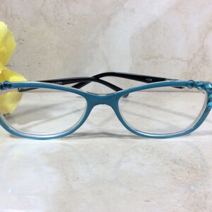 May include: Teal-colored reading glasses with black arms and clear lenses. The frames are adorned with blue rhinestones at the corners. The glasses have a +2.00 magnification. The text "CHINA" is printed on the arm.