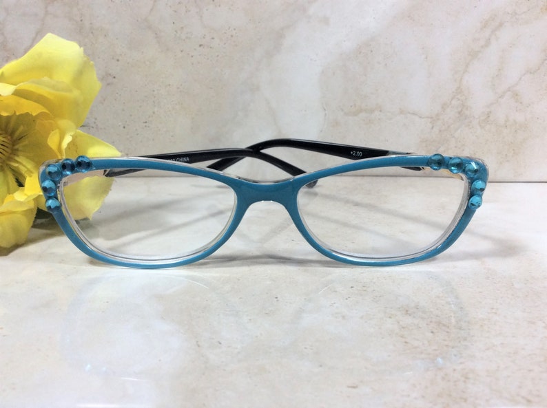 May include: Teal-colored reading glasses with black arms and clear lenses. The frames are adorned with blue rhinestones at the corners. The glasses have a +2.00 magnification. The text "CHINA" is printed on the arm.