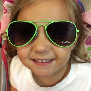 May include: A young girl wearing bright green sunglasses with the word "Hallie" printed on the right lens. The sunglasses have a lime green frame.
