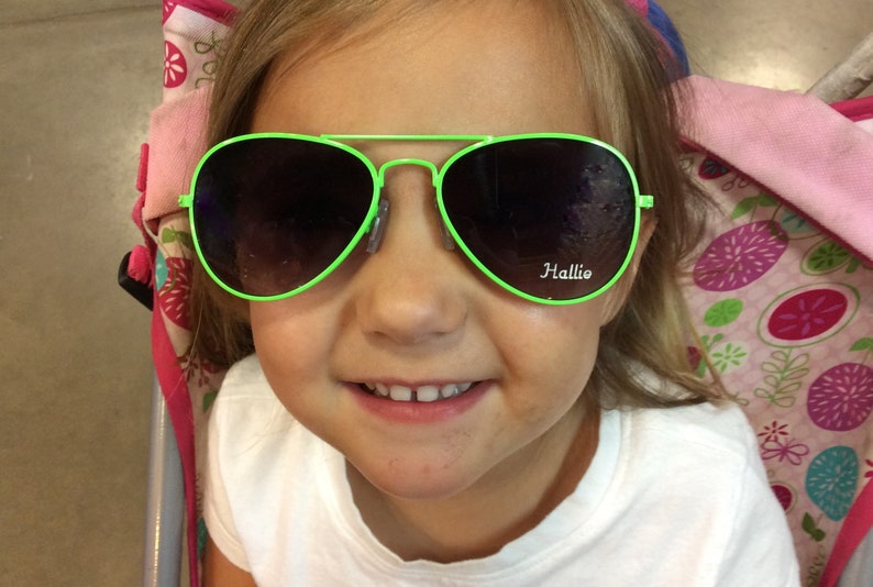 May include: A young girl wearing bright green sunglasses with the word "Hallie" printed on the right lens. The sunglasses have a lime green frame.