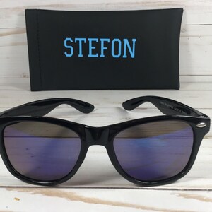 Pu&ograve; includere: Occhiali da sole neri con lenti a specchio blu. Gli occhiali da sole sono su una superficie di legno bianco. La custodia degli occhiali da sole &egrave; nera con la parola "STEFON" stampata in blu.