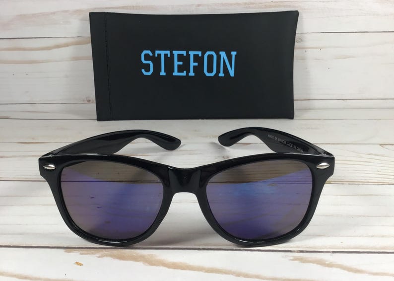 Pu&ograve; includere: Occhiali da sole neri con lenti a specchio blu. Gli occhiali da sole sono su una superficie di legno bianco. La custodia degli occhiali da sole &egrave; nera con la parola "STEFON" stampata in blu.
