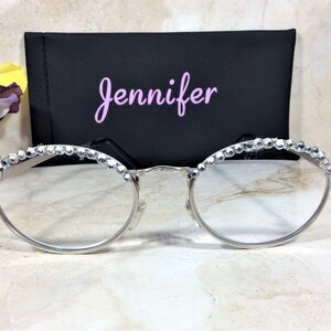 May include: Silver-rimmed eyeglasses with clear lenses, adorned with sparkling rhinestones along the top frame. A black case with the name "Jennifer" in pink script is behind the glasses. The overall aesthetic is elegant and stylish.