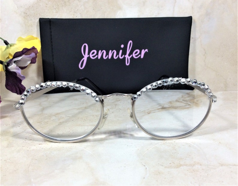 May include: Silver-rimmed eyeglasses with clear lenses, adorned with sparkling rhinestones along the top frame. A black case with the name "Jennifer" in pink script is behind the glasses. The overall aesthetic is elegant and stylish.