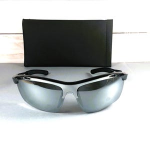 May include: Sport sunglasses featuring a sleek silver mirrored lens and a black frame with a wrap-around design. The eyewear is accompanied by a rectangular black protective case with a smooth finish, positioned directly behind the glasses.