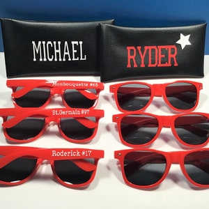 May include: Red sunglasses with dark lenses are displayed, some with names and numbers printed on the frames. Two black cases are visible, one with "MICHAEL" and the other with "RYDER" and a white star.
