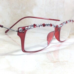 May include: Red-framed eyeglasses with clear lenses. The top of the frames are embellished with a row of clear and red rhinestones. The temples are a thin, silver metal. The glasses are set against a light-colored background.