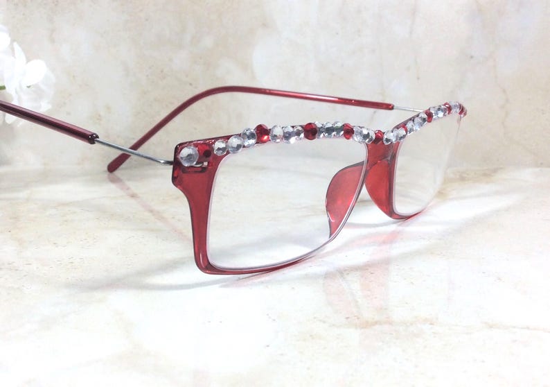 May include: Red-framed eyeglasses with clear lenses. The top of the frames are embellished with a row of clear and red rhinestones. The temples are a thin, silver metal. The glasses are set against a light-colored background.