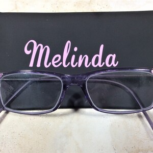 May include: Purple rectangular eyeglasses with clear lenses and pink and white rhinestones on the outer corners. The glasses are positioned in front of a black rectangular case with the name Melinda printed in light pink script lettering.