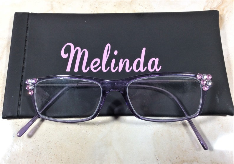 May include: Purple rectangular eyeglasses with clear lenses and pink and white rhinestones on the outer corners. The glasses are positioned in front of a black rectangular case with the name Melinda printed in light pink script lettering.