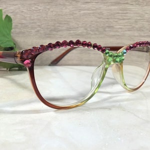May include: A pair of eyeglasses with a gradient brown and green frame. The top of the frame is adorned with pink rhinestones. A cluster of green and blue rhinestones decorates the bridge of the glasses.