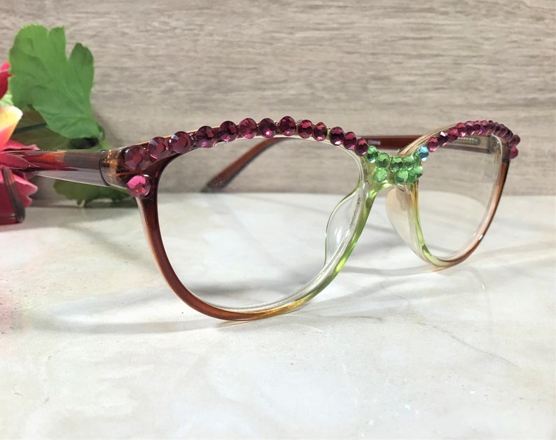 May include: A pair of eyeglasses with a gradient brown and green frame. The top of the frame is adorned with pink rhinestones. A cluster of green and blue rhinestones decorates the bridge of the glasses.