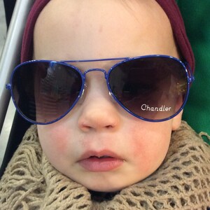 May include: A baby wearing a blue and brown scarf and blue aviator sunglasses with the word "Chandler" on the right lens.