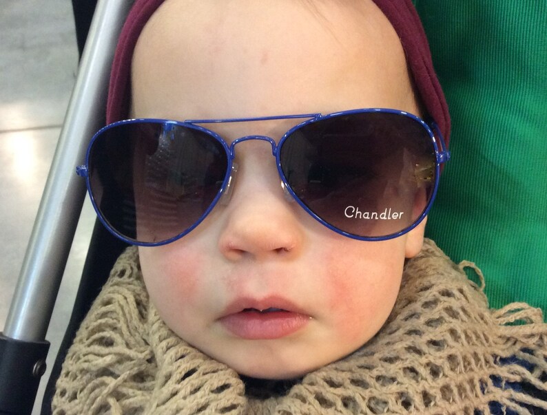 May include: A baby wearing a blue and brown scarf and blue aviator sunglasses with the word "Chandler" on the right lens.