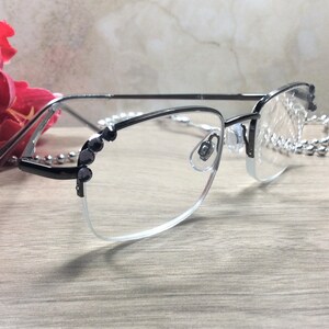 May include: Black and silver-toned eyeglasses with a half-rim design. The frames feature decorative black accents on the corners and temples. A silver-toned chain is draped near the glasses. A red flower is in the background.