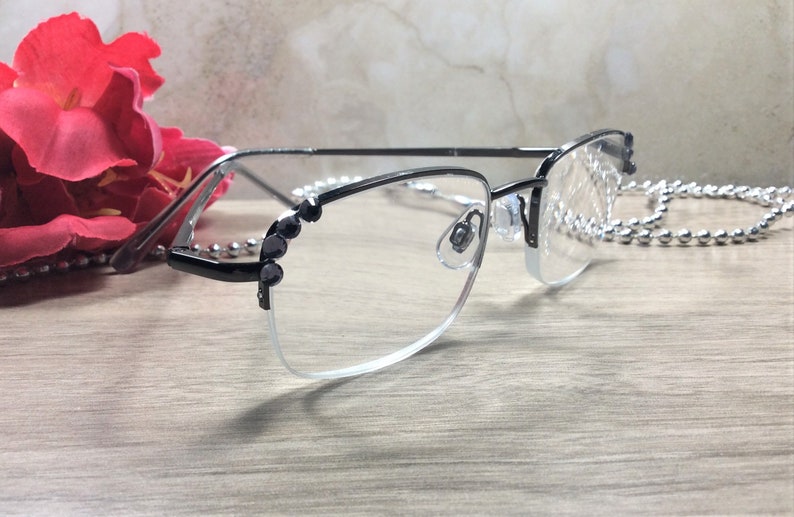 May include: Black and silver-toned eyeglasses with a half-rim design. The frames feature decorative black accents on the corners and temples. A silver-toned chain is draped near the glasses. A red flower is in the background.