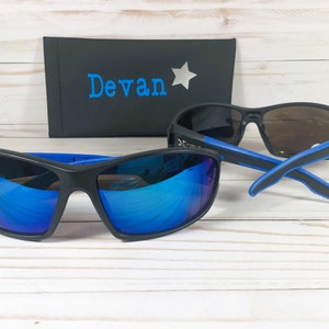 May include: Two pairs of black sunglasses with blue accents. The sunglasses have mirrored lenses. One pair has blue mirrored lenses and the other pair has brown mirrored lenses. The sunglasses are on a white wooden surface. A black case with the name "Devan" and a silver star is in the background.
