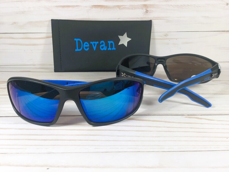May include: Two pairs of black sunglasses with blue accents. The sunglasses have mirrored lenses. One pair has blue mirrored lenses and the other pair has brown mirrored lenses. The sunglasses are on a white wooden surface. A black case with the name "Devan" and a silver star is in the background.