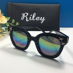 Puede incluir: Gafas de sol negras de ojo de gato con lentes iridiscentes y detalles de estrellas doradas. Las gafas est&aacute;n en una funda negra con la palabra "Riley" impresa en ella.