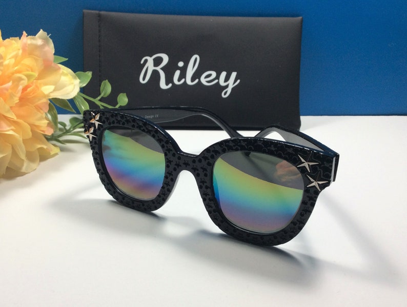 Puede incluir: Gafas de sol negras de ojo de gato con lentes iridiscentes y detalles de estrellas doradas. Las gafas est&aacute;n en una funda negra con la palabra "Riley" impresa en ella.
