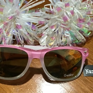 May include: Pink sunglasses with a clear frame and mirrored lenses. The sunglasses have the name "Gabby" engraved on the right lens. A tag is attached to the sunglasses that says "Name Engraved FREE".