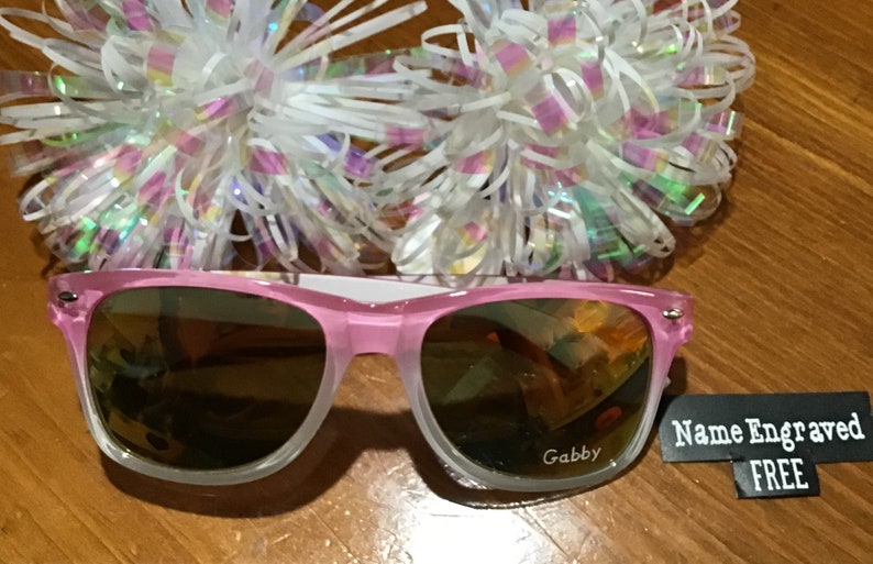 May include: Pink sunglasses with a clear frame and mirrored lenses. The sunglasses have the name "Gabby" engraved on the right lens. A tag is attached to the sunglasses that says "Name Engraved FREE".