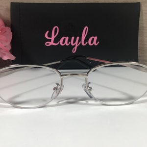 K&ouml;nnte beinhalten: Brille mit silbernem Rahmen, klaren Gl&auml;sern und dekorativen Strasssteinen an den B&uuml;geln. Die Brille befindet sich vor einem schwarzen Etui mit dem Namen "Layla" in rosa Schrift. Eine rosa Blume ist im Hintergrund.