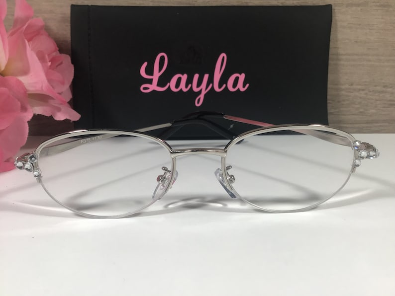 K&ouml;nnte beinhalten: Brille mit silbernem Rahmen, klaren Gl&auml;sern und dekorativen Strasssteinen an den B&uuml;geln. Die Brille befindet sich vor einem schwarzen Etui mit dem Namen "Layla" in rosa Schrift. Eine rosa Blume ist im Hintergrund.