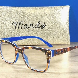 Puede incluir: Gafas con montura de carey y detalles azules. Las monturas est&aacute;n adornadas con tachuelas doradas. En el fondo, un estuche dorado con lentejuelas con el nombre "Mandy" en escritura cursiva.