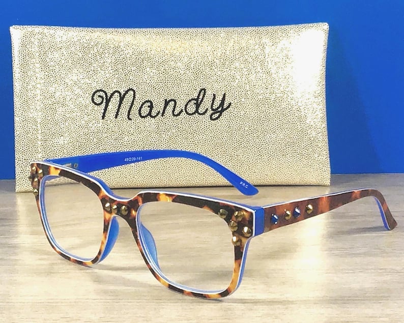 Puede incluir: Gafas con montura de carey y detalles azules. Las monturas est&aacute;n adornadas con tachuelas doradas. En el fondo, un estuche dorado con lentejuelas con el nombre "Mandy" en escritura cursiva.