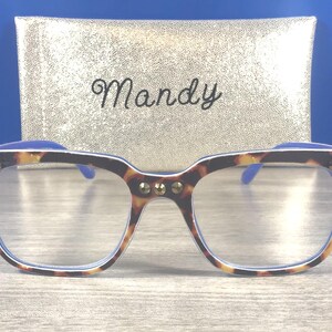 Puede incluir: Gafas de montura cuadrada con estampado de carey y tachuelas doradas. Se ven las patillas y lentes azules. Detr&aacute;s de las gafas hay un estuche dorado con el nombre "Mandy" en cursiva.