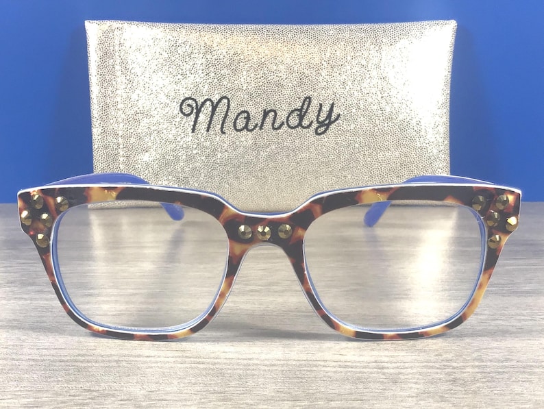 Puede incluir: Gafas de montura cuadrada con estampado de carey y tachuelas doradas. Se ven las patillas y lentes azules. Detr&aacute;s de las gafas hay un estuche dorado con el nombre "Mandy" en cursiva.