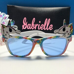May include: Rainbow unicorn sunglasses with blue lenses and a white frame. The sunglasses have a unicorn head on each side with pink, yellow, and blue details. The name "Gabrielle" is written on a black case behind the sunglasses.