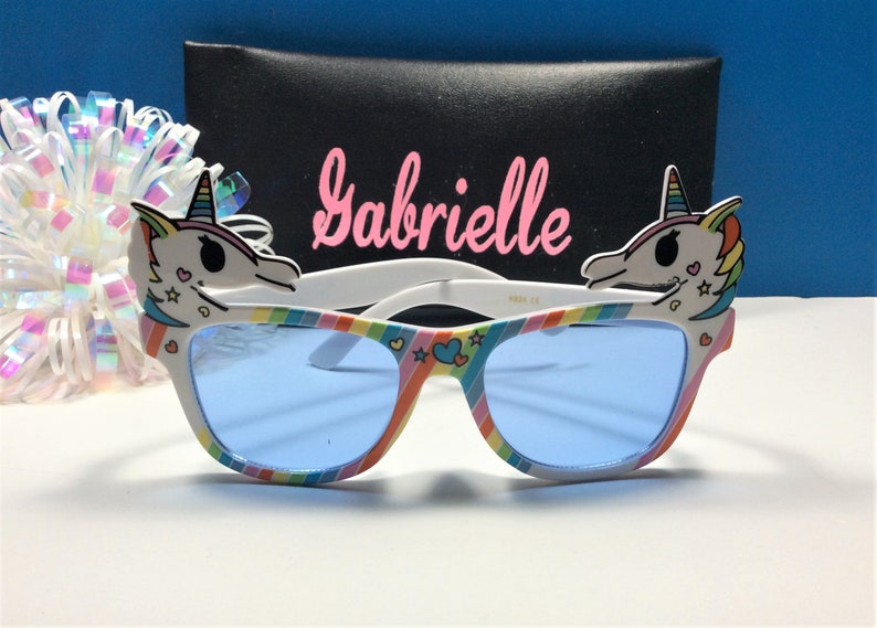 May include: Rainbow unicorn sunglasses with blue lenses and a white frame. The sunglasses have a unicorn head on each side with pink, yellow, and blue details. The name "Gabrielle" is written on a black case behind the sunglasses.