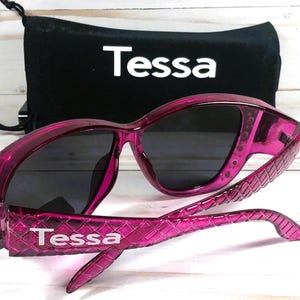 May include: Pink sunglasses with dark lenses and a matching black drawstring pouch. The sunglasses have a textured pattern on the arms and the brand name "Tessa" is printed on the arms and pouch.