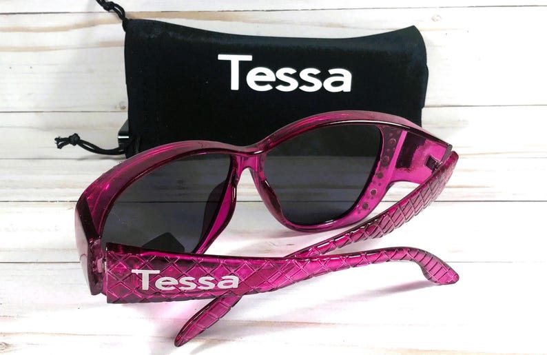 May include: Pink sunglasses with dark lenses and a matching black drawstring pouch. The sunglasses have a textured pattern on the arms and the brand name "Tessa" is printed on the arms and pouch.