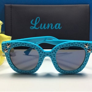 Puede incluir: Gafas de sol de color azul turquesa con detalles de estrellas plateadas. Las gafas tienen una superficie texturizada y est&aacute;n en una caja con la palabra "Luna" impresa en ella.