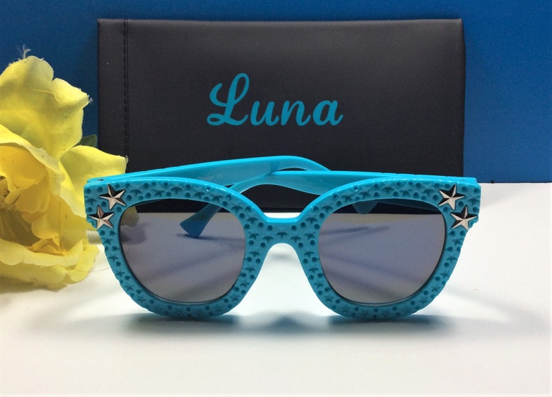 Puede incluir: Gafas de sol de color azul turquesa con detalles de estrellas plateadas. Las gafas tienen una superficie texturizada y est&aacute;n en una caja con la palabra "Luna" impresa en ella.