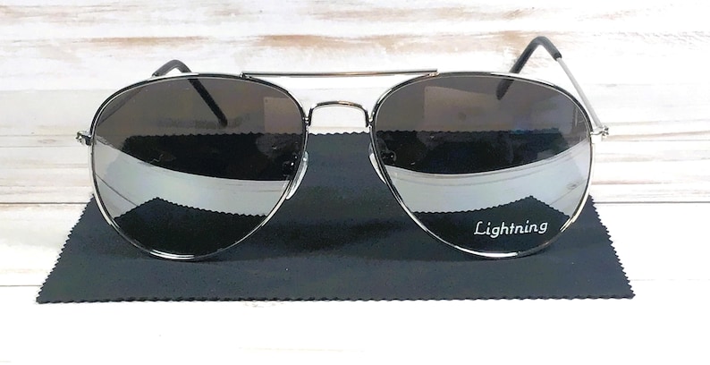 May include: Aviator-style sunglasses with dark lenses and a silver frame. The sunglasses rest on a black microfiber cloth. The text "Lightning" is printed on the lens.
