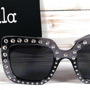 May include: Black square sunglasses with rhinestones around the frame. The sunglasses have dark lenses.