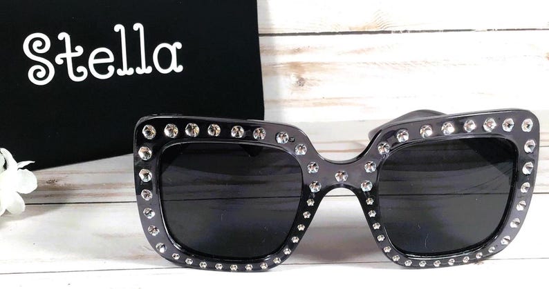 May include: Black square sunglasses with rhinestones around the frame. The sunglasses have dark lenses.