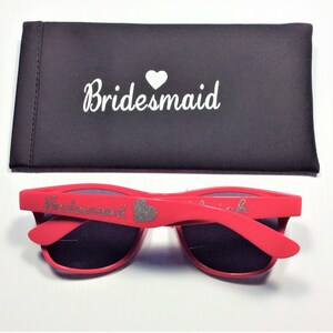 May include: Red sunglasses with a silver glitter heart and the word "Bridemaid" on the frame. The sunglasses come with a black carrying case that says "Bridemaid" with a white heart.