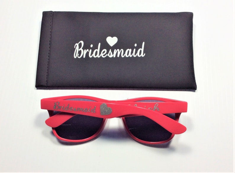 May include: Red sunglasses with a silver glitter heart and the word "Bridemaid" on the frame. The sunglasses come with a black carrying case that says "Bridemaid" with a white heart.