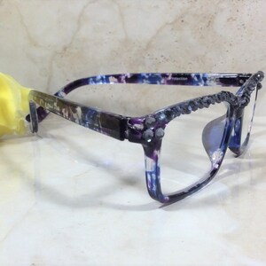 Op de afbeelding: Bril met een paars en blauw gemarmerd patroon op het montuur. De bovenranden aan de voorzijde zijn versierd met een rij kleine, donkergrijze gefacetteerde kristallen. De pootjes hebben een bijpassend marmerontwerp en binnenin staat UV Protection.