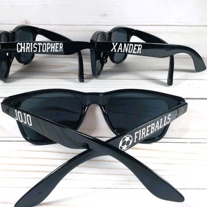 May include: Three pairs of black sunglasses with white text. The sunglasses on the top have the names "Christopher" and "Xander" on them. The sunglasses on the bottom have the name "Jojo" and the words "Fireballs" with a soccer ball graphic.
