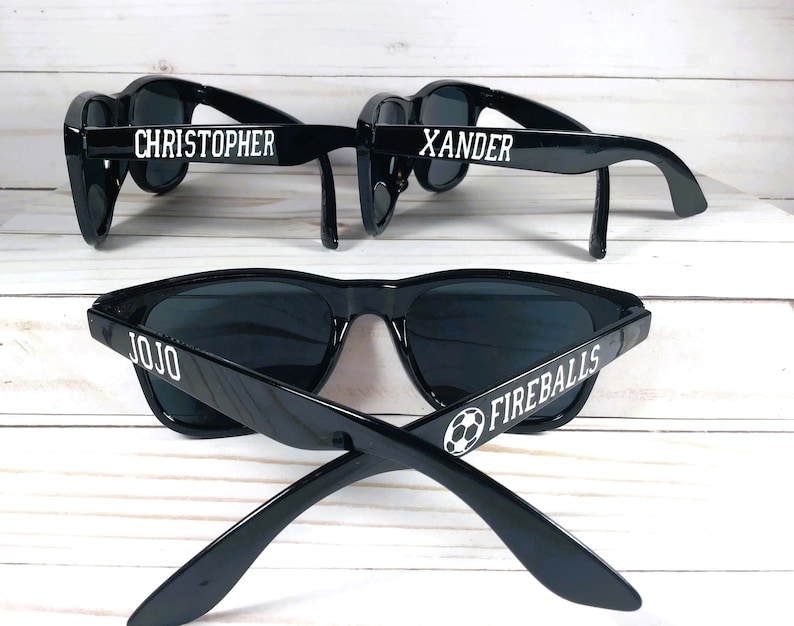 May include: Three pairs of black sunglasses with white text. The sunglasses on the top have the names "Christopher" and "Xander" on them. The sunglasses on the bottom have the name "Jojo" and the words "Fireballs" with a soccer ball graphic.