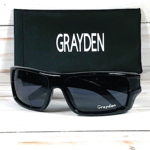May include: Black sunglasses with a matching black case. The sunglasses have a thick frame and dark lenses. The case is rectangular and features the name "GRAYDEN" in white block letters. The brand name "Grayden" is also printed on the sunglasses.