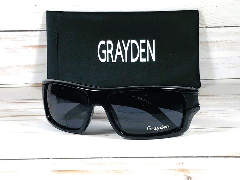 May include: Black sunglasses with a matching black case. The sunglasses have a thick frame and dark lenses. The case is rectangular and features the name "GRAYDEN" in white block letters. The brand name "Grayden" is also printed on the sunglasses.